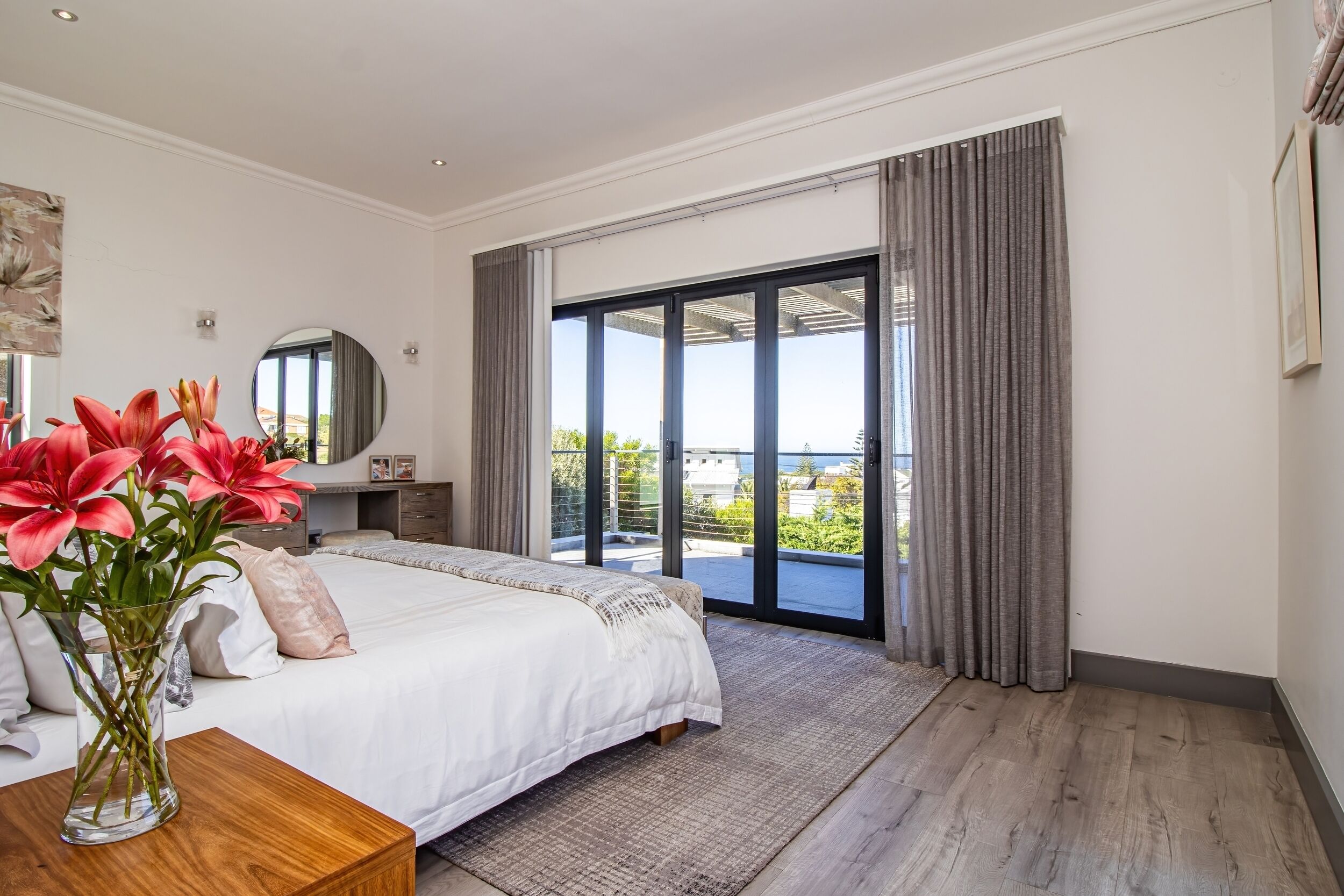 A bedroom with a large bed, a nightstand, a vase with flowers, a mirror, and a view of the outdoors through a sliding glass door.