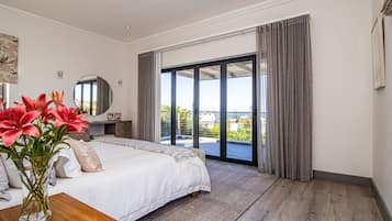 A bedroom with a large bed, a nightstand, a vase with flowers, a mirror, and a view of the outdoors through a sliding glass door.