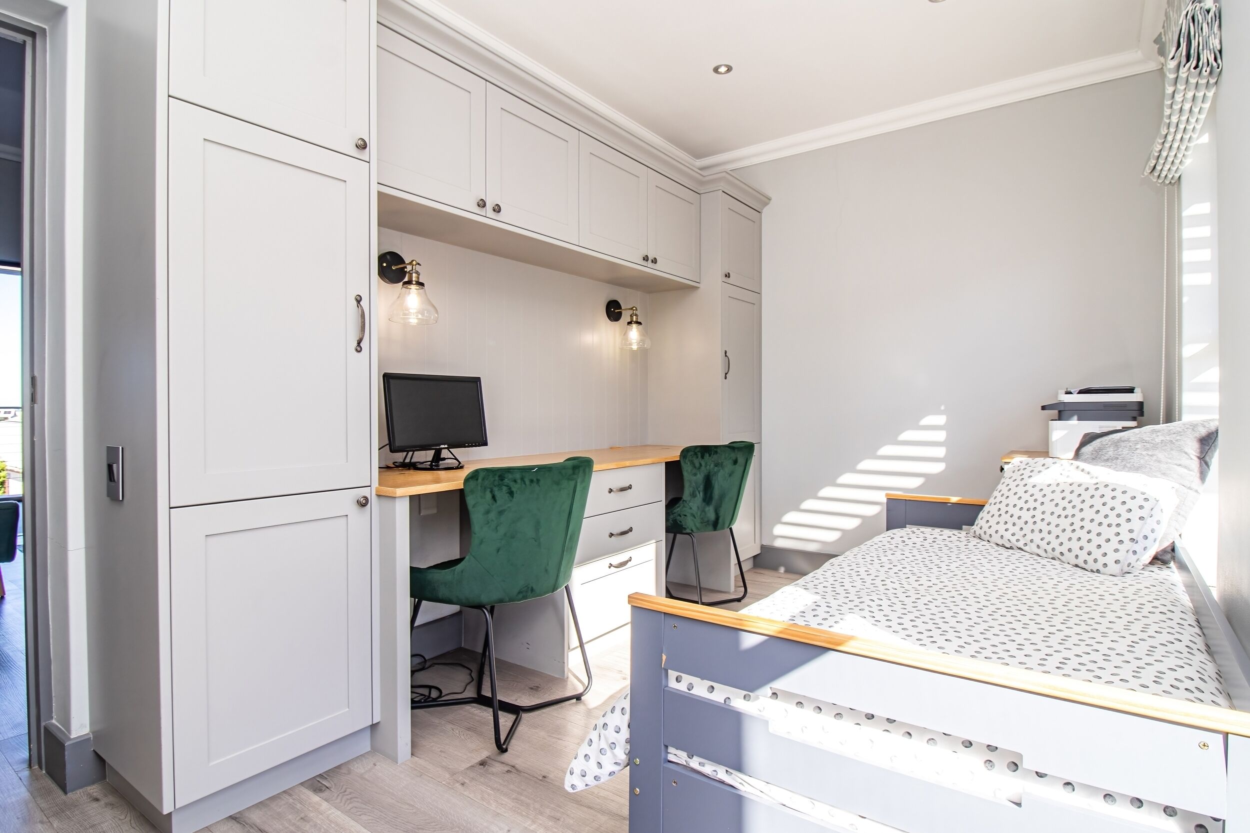 A bedroom with a bed, a desk with a computer, two green chairs, and a wardrobe.
