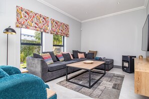 Una sala de estar moderna con un sofá gris, sillones turquesa, una mesa de centro de madera y un amplio ventanal con cortinas estampadas.