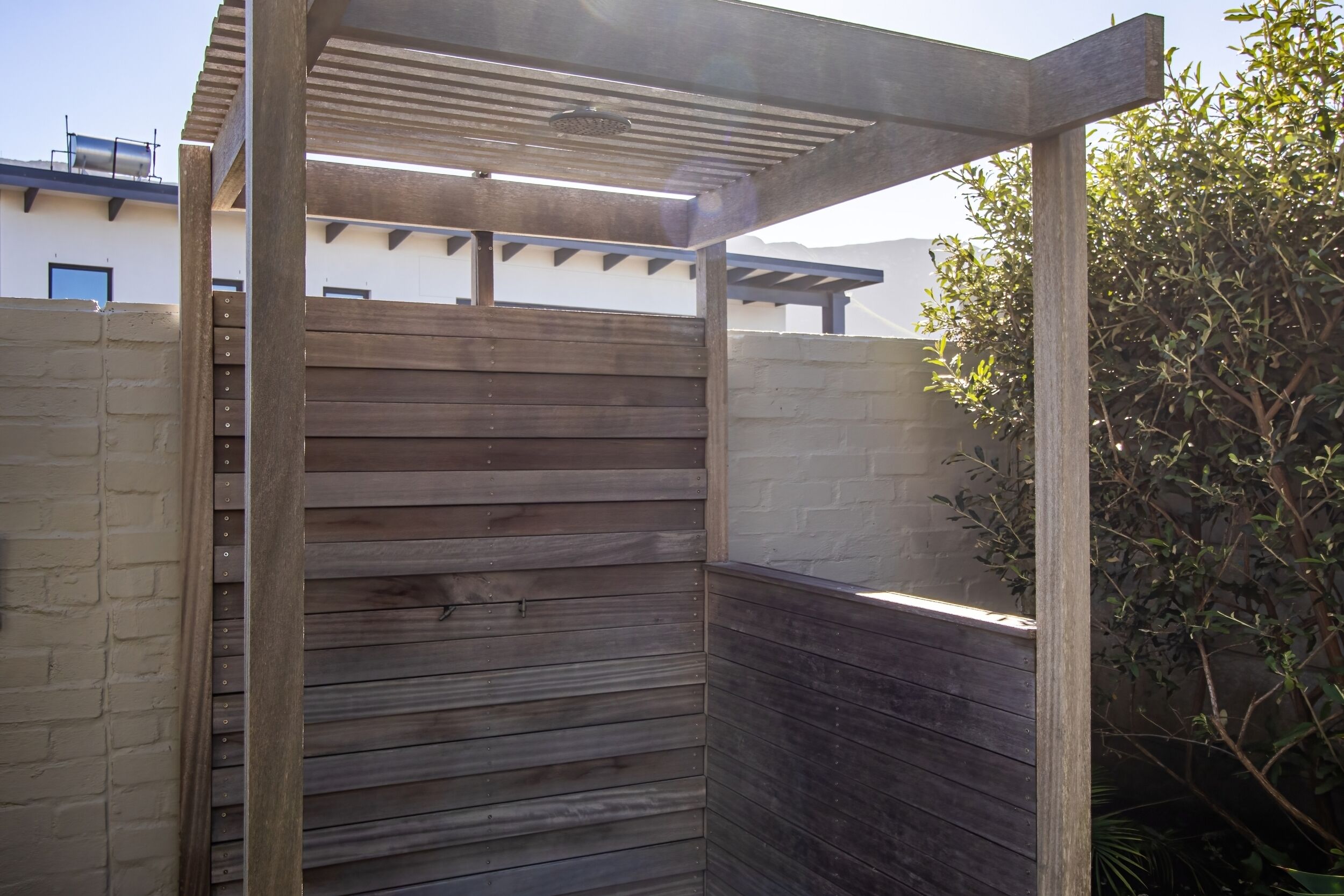 A wooden pergola structure with a metal roof, adjacent to a brick wall and a white building with a blue door.