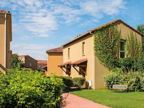 Un edificio residencial con techo de tejas, un porche cubierto y un jardín bien cuidado con un sendero y un banco.