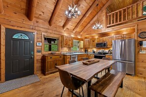 A wooden cabin interior with a dining table, chairs, a kitchen with appliances, and a large window. - Chimney Path on Fightingtown Creek - Pet Friendly | Creekside Firepit | Game Room (Mc Caysville)
