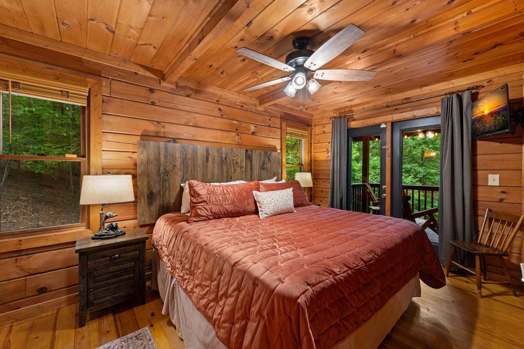 Dormitorio de cabaña de madera con cama, mesita de noche, lámpara, ventilador de techo y vistas al exterior.