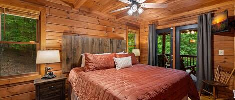 Dormitorio de cabaña de madera con cama, mesita de noche, lámpara, ventilador de techo y vistas al exterior.