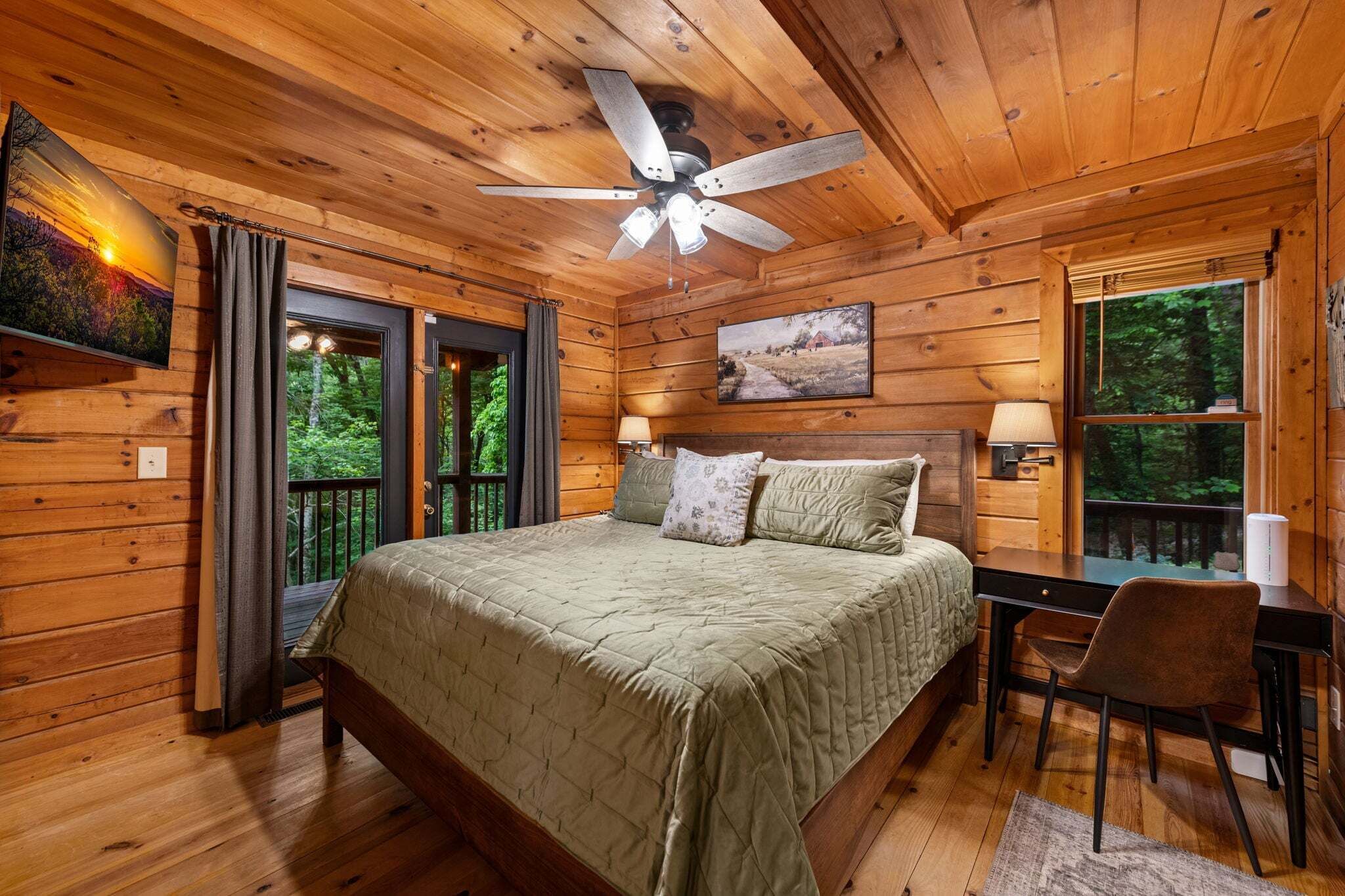 A wooden cabin bedroom with a bed, desk, chair, ceiling fan, and two windows.