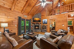 A log cabin interior with a fireplace, leather furniture, and a balcony with a view of greenery. - Chimney Path on Fightingtown Creek - Pet Friendly | Creekside Firepit | Game Room (Mc Caysville)