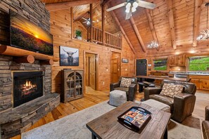 A log cabin interior with a stone fireplace, leather furniture, a television mounted on the wall, and a balcony. - Chimney Path on Fightingtown Creek - Pet Friendly | Creekside Firepit | Game Room (Mc Caysville)