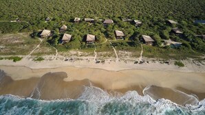 Ett flygbildsbild av en kustort med flera stugor med halmtak, en sandstrand och en bakgrund av tät grönska.