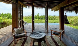 Une terrasse en bois avec un banc, une table et une chaise, offrant une vue sur un paysage verdoyant et luxuriant ainsi que sur la mer.