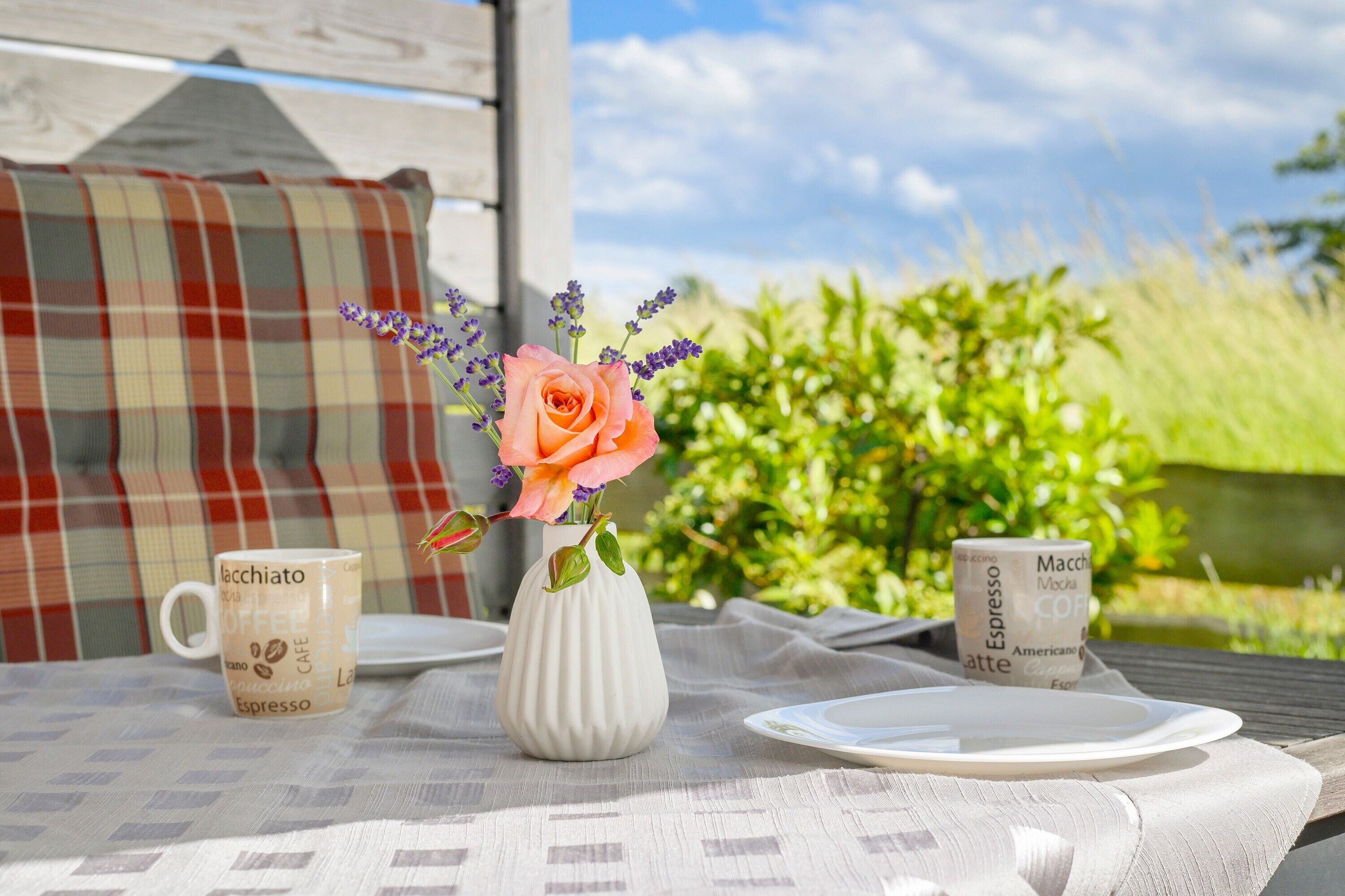 Une table sur laquelle se trouve un vase blanc contenant une rose et de la lavande, deux tasses à café et une assiette.