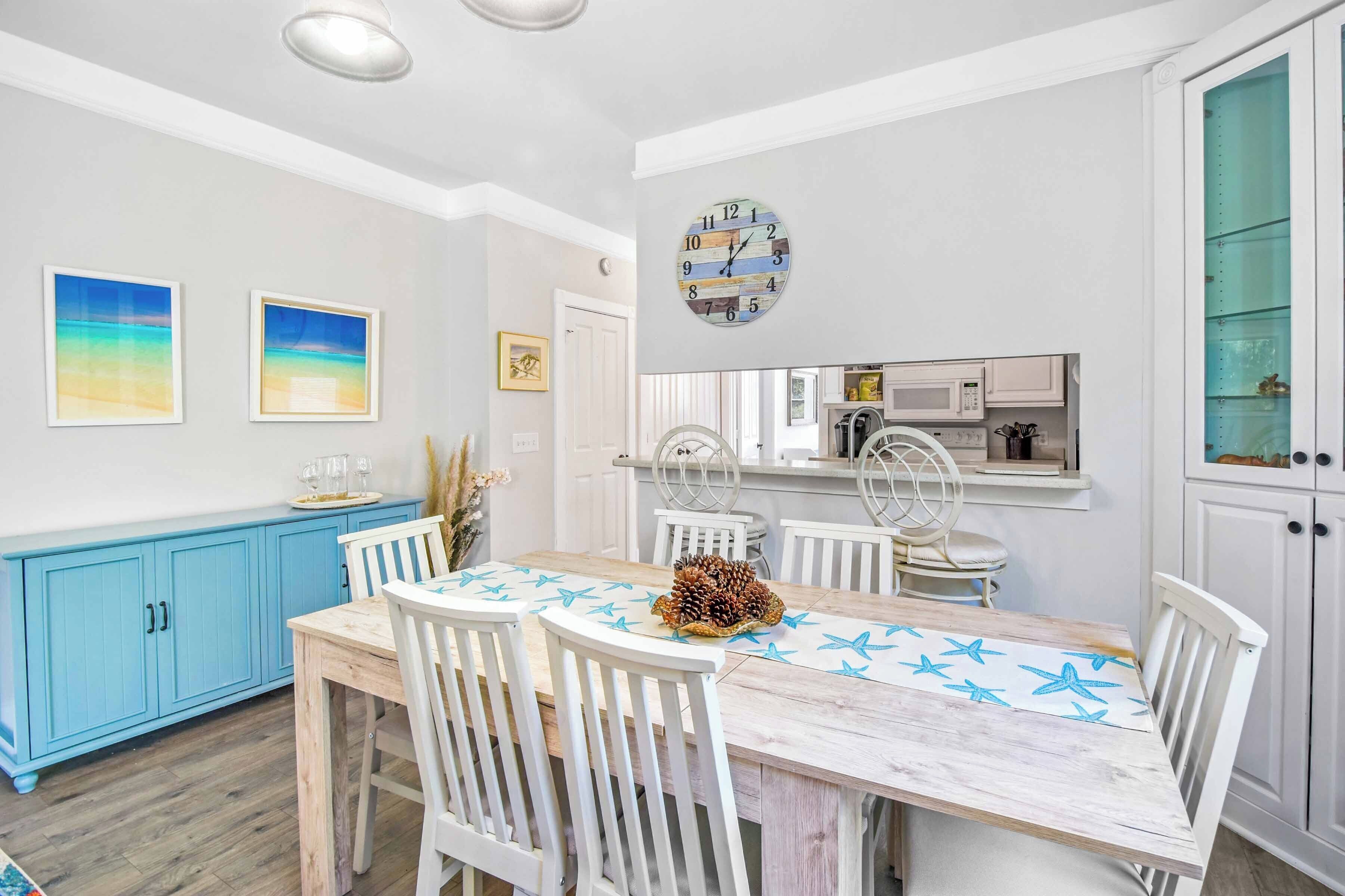 A dining area with a wooden table, white chairs, a clock, and a kitchen in the background.