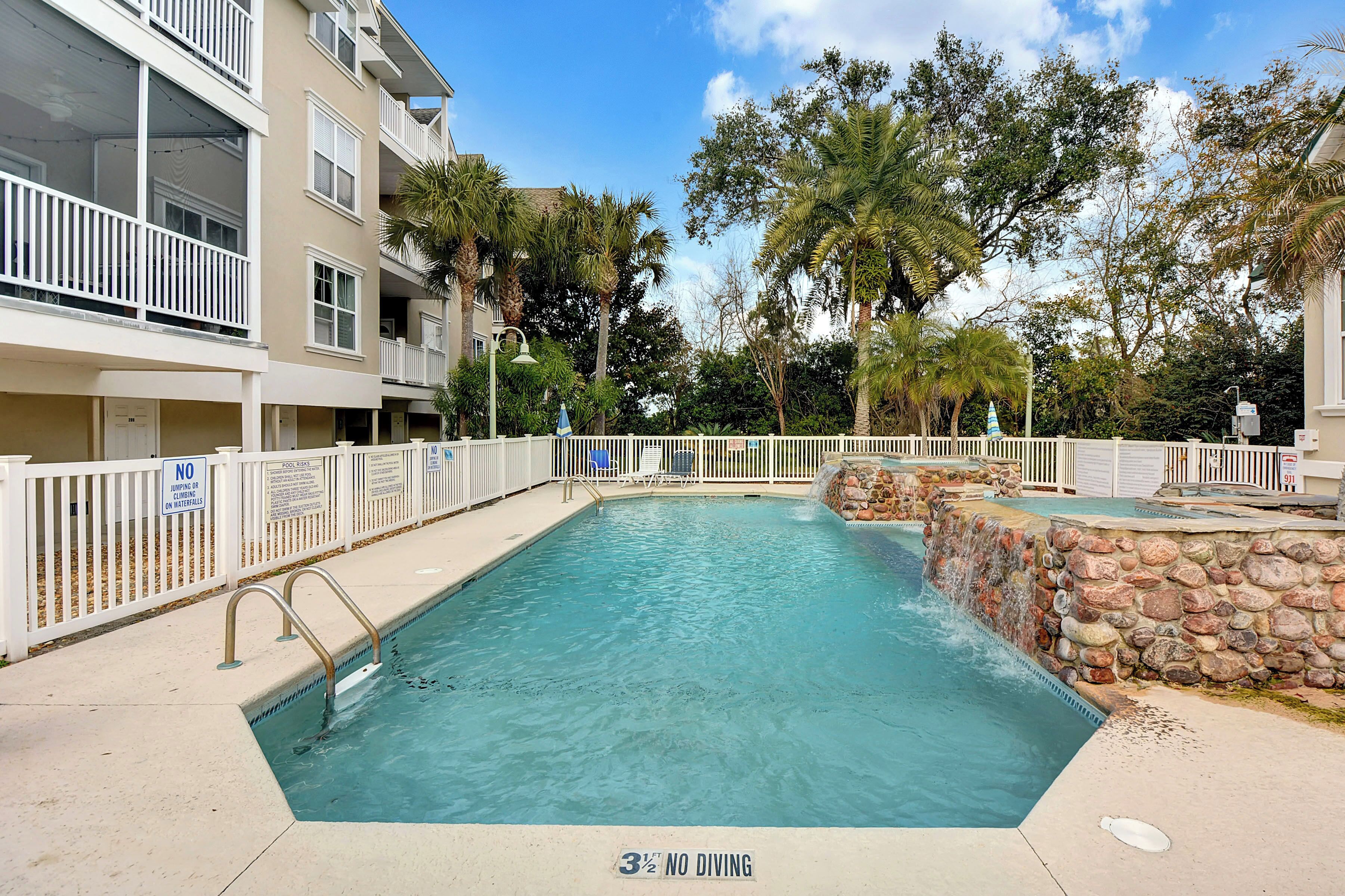 A swimming pool with a no diving sign, surrounded by a white fence, and a stone wall with a water feature.