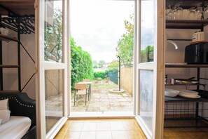 Una porta scorrevole aperta che dà su un patio con tavolo e sedie, un divano nero e un angolo cottura con scaffali e una macchina del caffè.