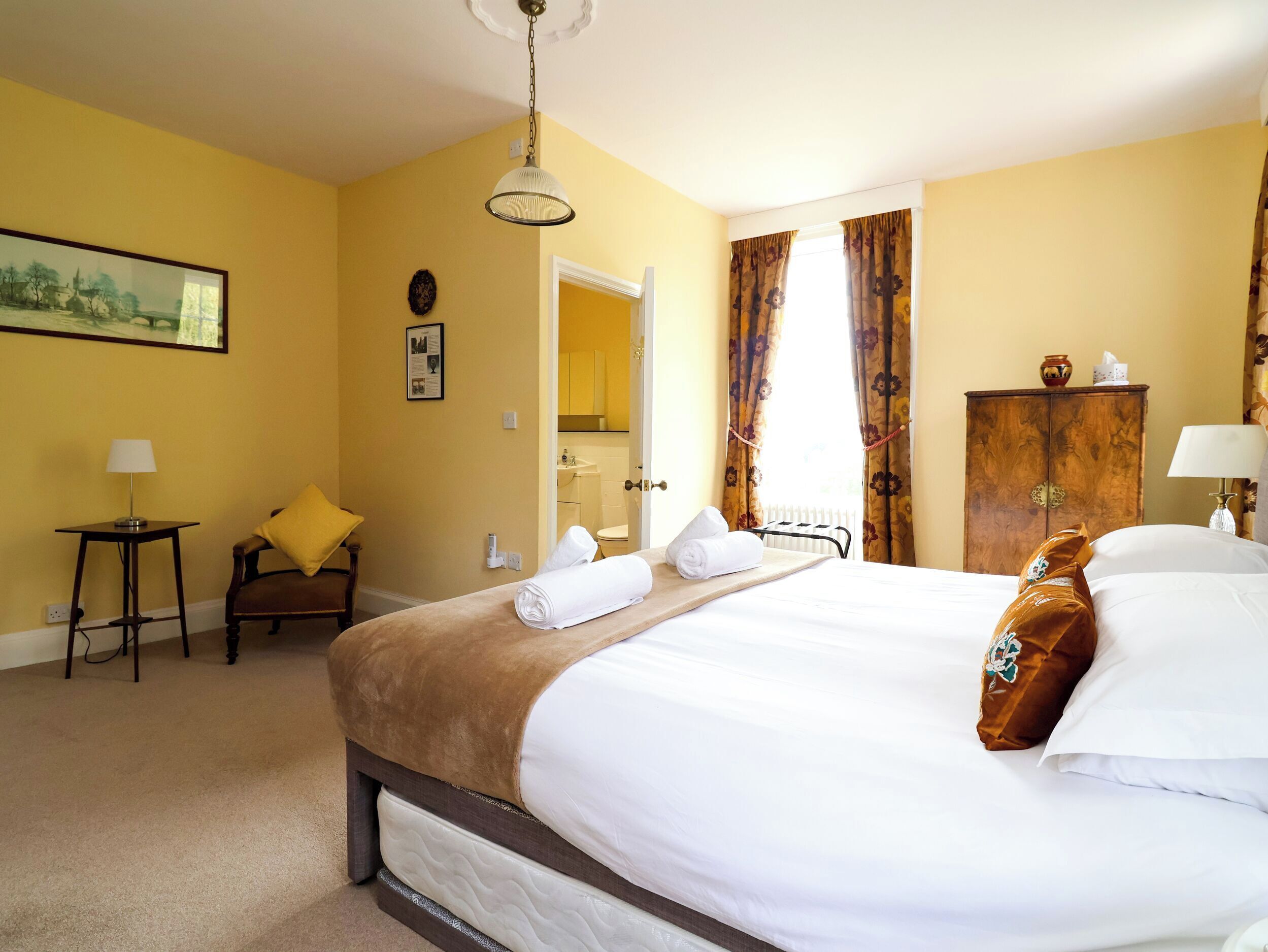 A hotel room with a bed, a chair, a small table, a lamp, a painting, and a wooden cabinet.