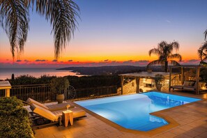 Un espace aménagé au bord de la piscine, offrant une vue sur le coucher de soleil sur la mer, avec des chaises longues et une pergola.