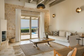 A spacious living room with a stone wall, a sofa, a coffee table, and a view of the sea. - Villa Ainos of Lithos Villas (Vlachata)