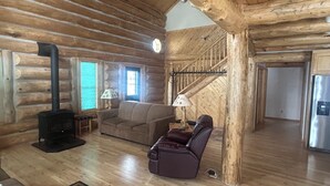 A log cabin interior with a fireplace, sofa, armchair, and a staircase. - Cracked Noggin Lodge (Dubois)