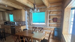 A wooden cabin interior with a dining table, chairs, a chandelier, a kitchen area, and a window with blinds. - Cracked Noggin Lodge (Dubois)