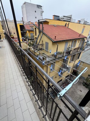 Een balkon met een metalen hekwerk, betegelde vloer en uitzicht op omliggende gebouwen met balkons en rode dakpannen.