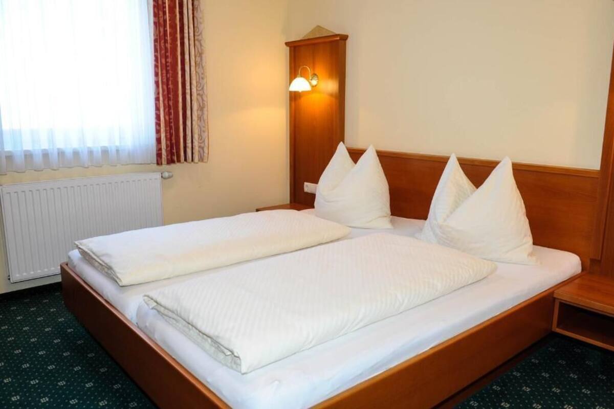 A double bed with white bedding, a wooden headboard, a nightstand, a wall-mounted light, and a window with curtains.