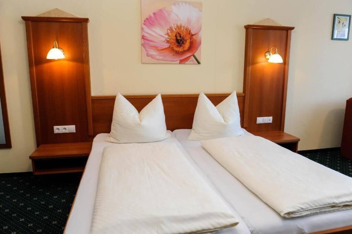 A hotel room with two beds, wooden headboards, bedside lamps, a wall-mounted flower painting, and a small nightstand.