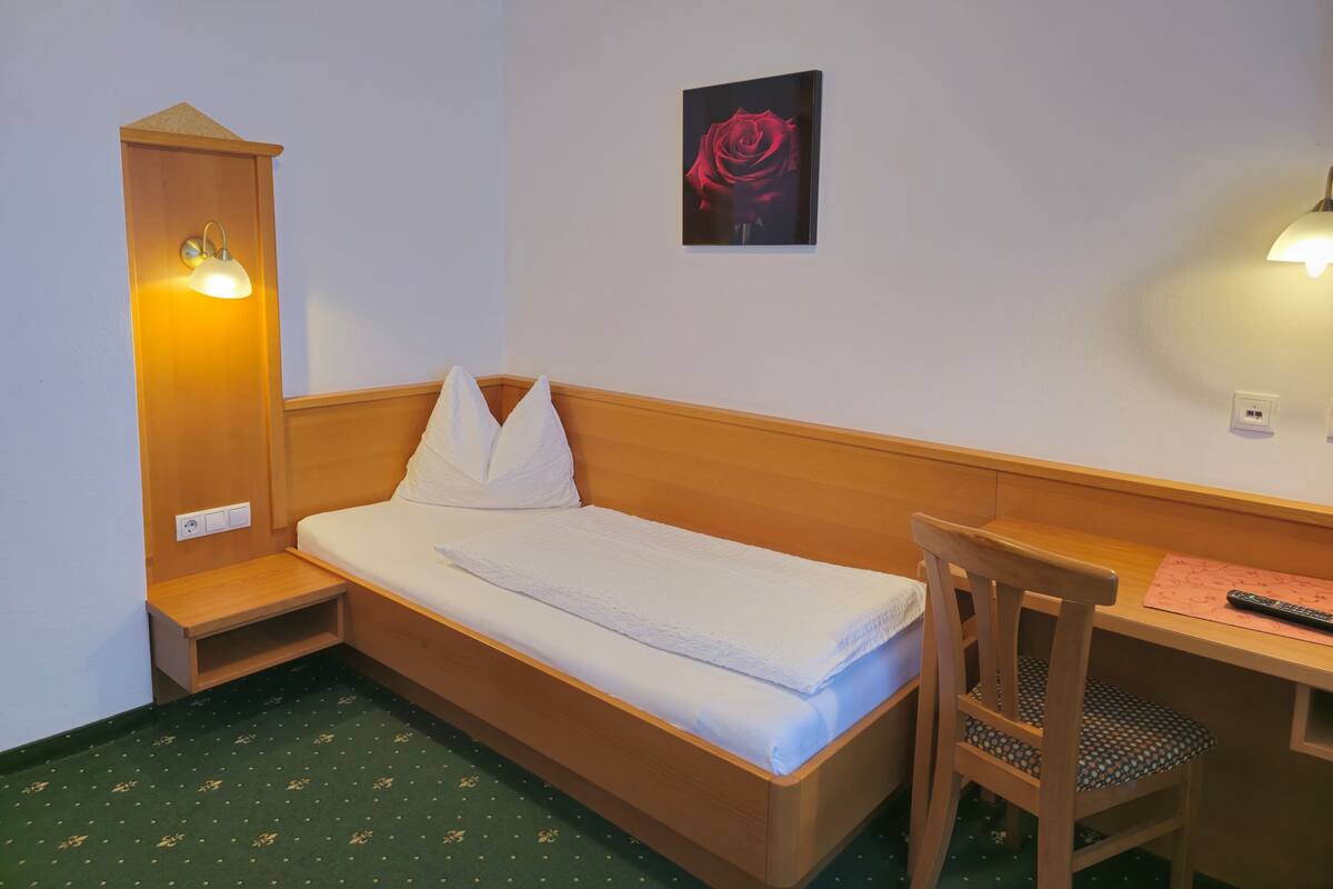 A single bed with a wooden headboard, a small wooden desk with a chair, a wall-mounted light, and a framed picture of a rose.