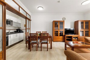 Eine moderne Küche mit weißen Schränken, einer Mikrowelle und einer Kaffeemaschine. Es gibt eine Essecke mit Holztischen und -stühlen. Im Wohnzimmer steht ein Fernseher auf einem Schrank.