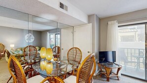 Een hotelkamer met een glazen eettafel, rieten stoelen, een flatscreen televisie, een balkon met uitzicht en een schuifdeur van glas.