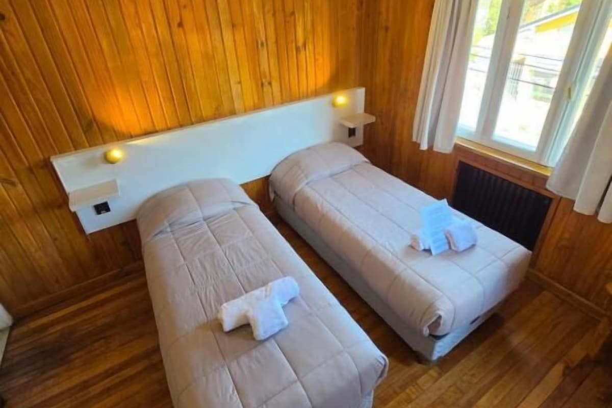 Habitación con paredes de madera, dos camas con sábanas blancas, una mesita de noche y una ventana con cortinas.
