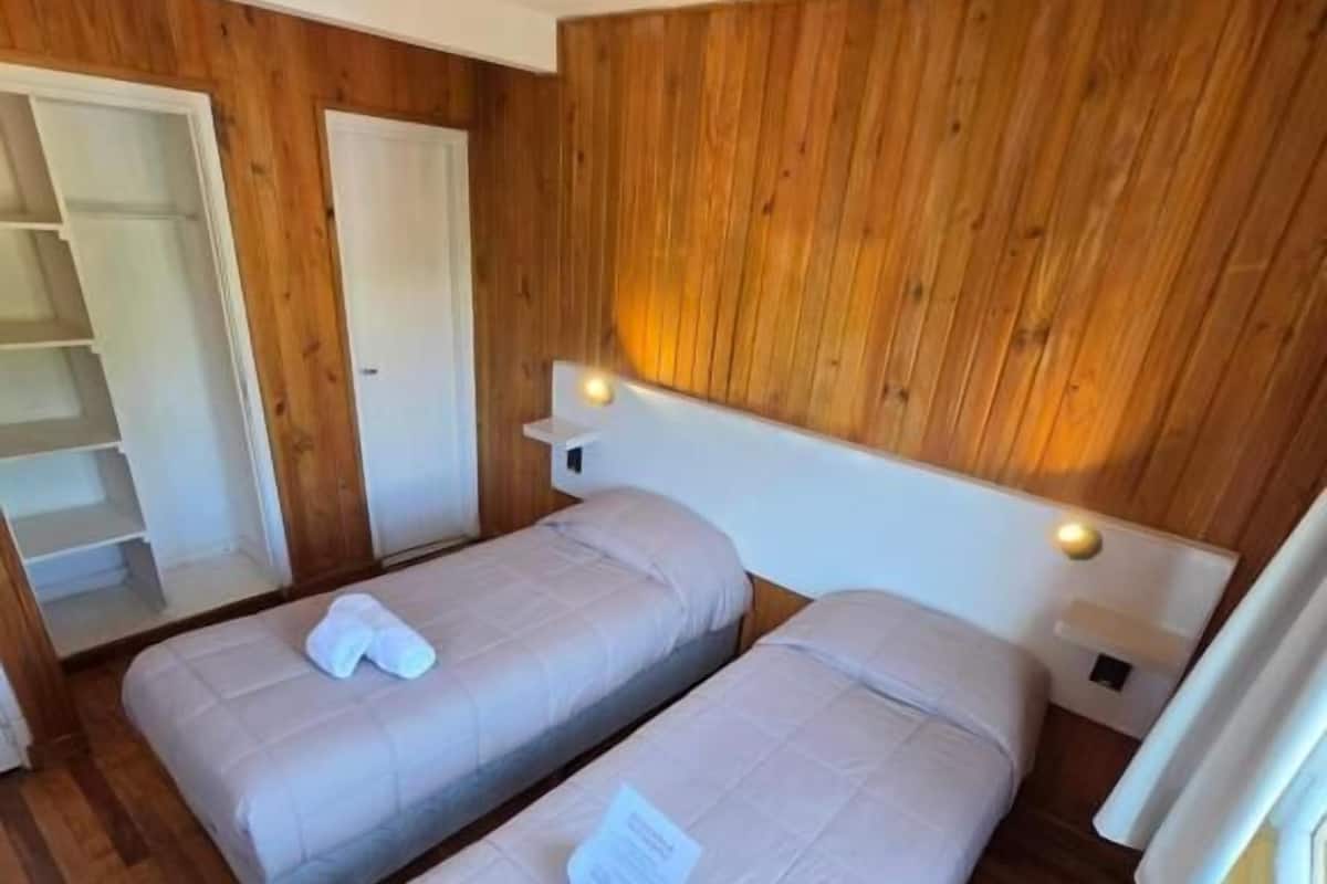 A room with two beds, wooden walls, a wardrobe, and a small shelf.
