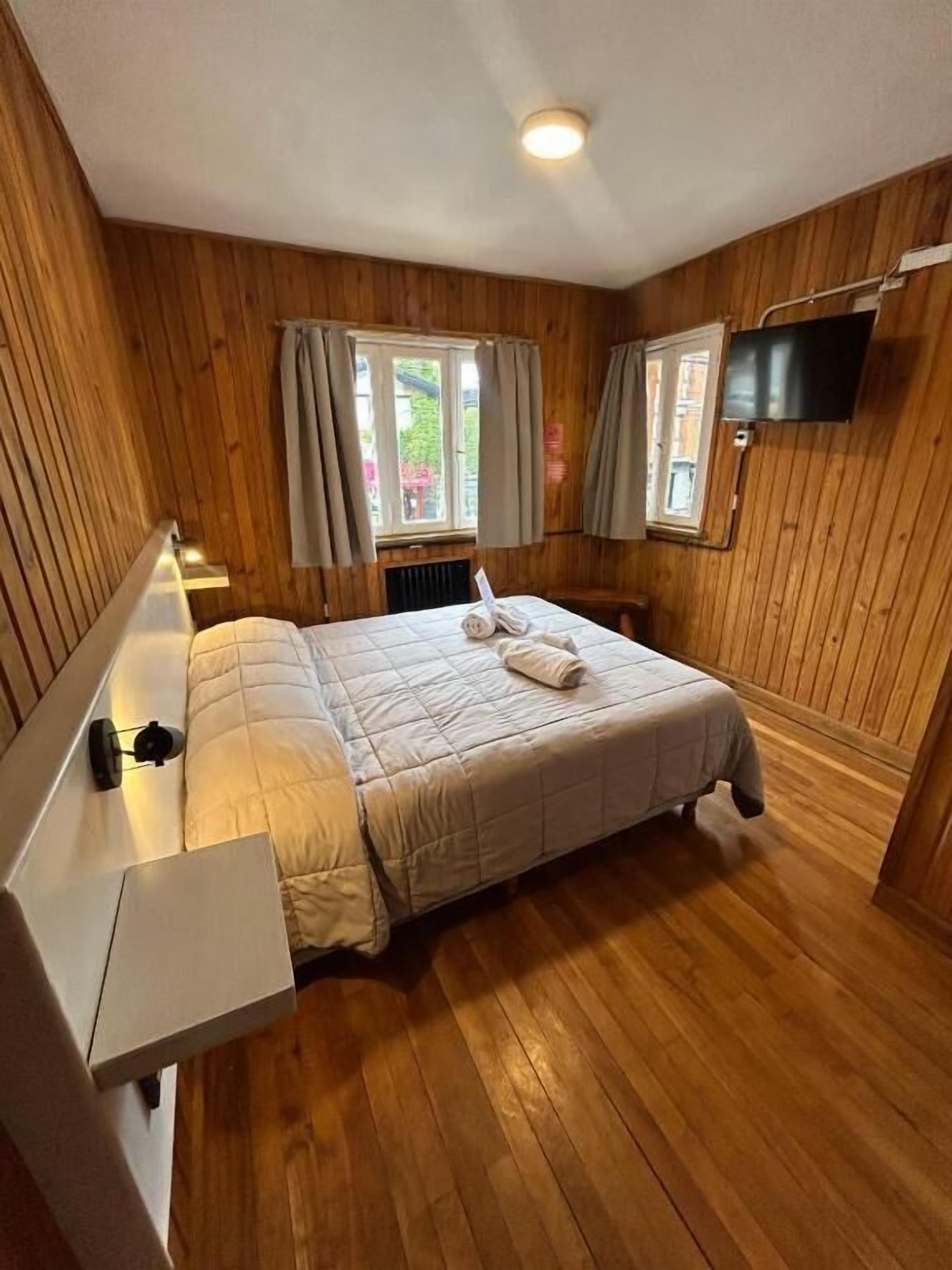 Ein Zimmer mit Holzwänden, einem Bett, einem an der Wand befestigten Fernseher und einem Fenster mit Vorhängen.