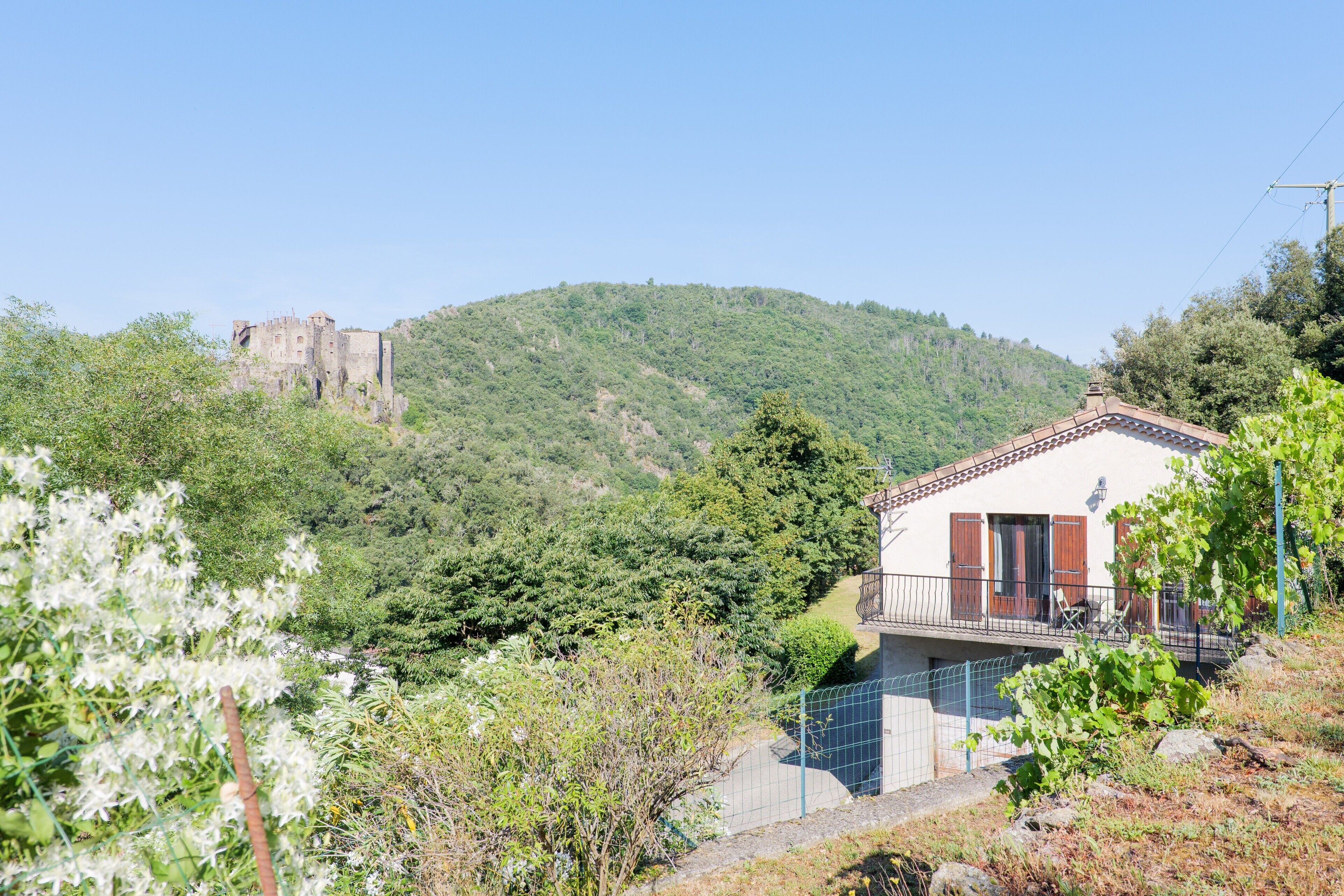 Une maison dotée d’un balcon, entourée de verdure, avec une colline en arrière-plan sur laquelle se dresse un château.