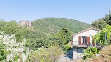 Une maison dotée d’un balcon, entourée de verdure, avec une colline en arrière-plan sur laquelle se dresse un château.
