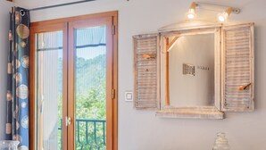 Une pièce dotée d’une porte-fenêtre coulissante, d’une commode en bois, d’un miroir à volets et d’une petite table sur laquelle se trouve un pot.
