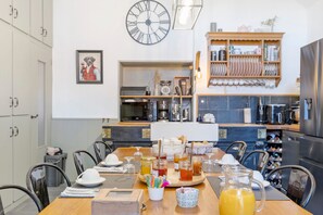 Un coin repas avec une table en bois dressée pour quatre personnes, une grande horloge murale et un espace cuisine équipé de divers appareils et d’un meuble à bouteilles.