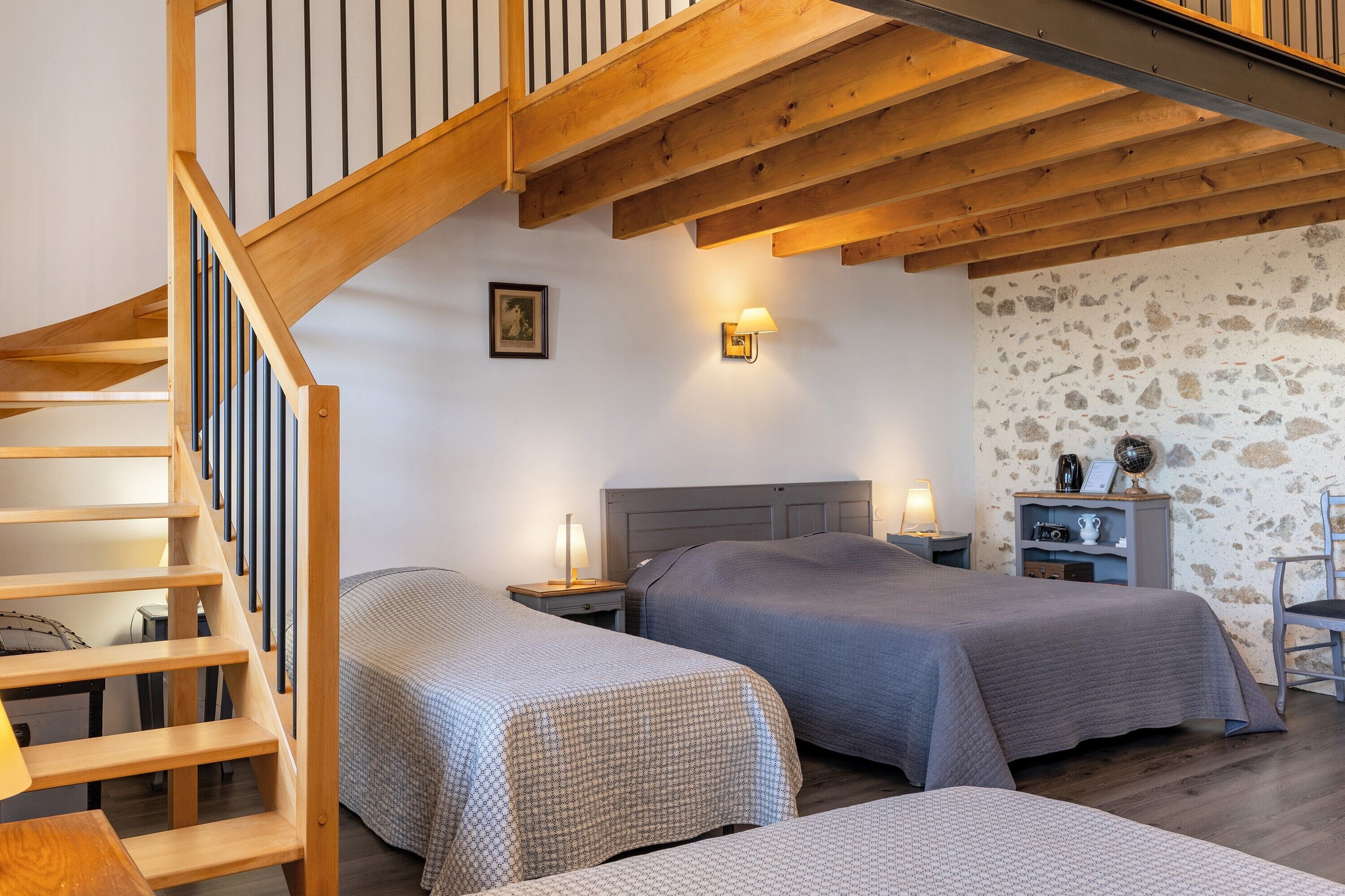 Ein Zimmer mit einer Holztreppe, zwei Betten mit grauer Bettwäsche, einem Nachttisch mit einer Lampe und einer Steinwand.
