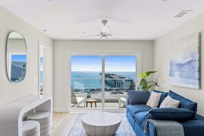 Ein modernes Wohnzimmer mit einem großen Fenster mit Blick auf einen Steg und den Ozean, einem Deckenventilator und einem blauen Sofa.