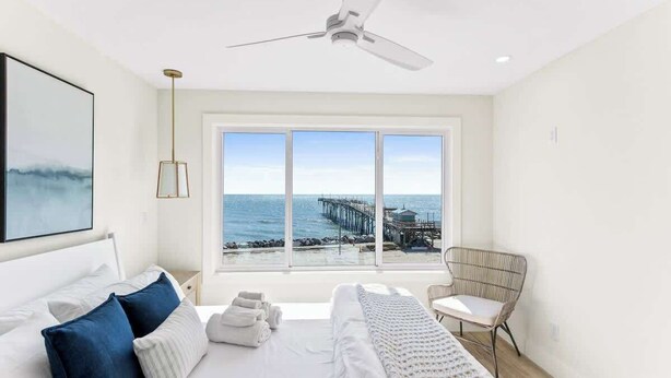 A bedroom with a bed, a chair, a ceiling fan, and a large window with a view of the ocean and pier. - Seaside Tranquility|North Pier Ocean Villa+Pool| (Carolina Beach)