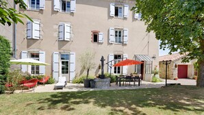 A building with multiple windows, a patio with outdoor furniture, and a tree providing shade.
