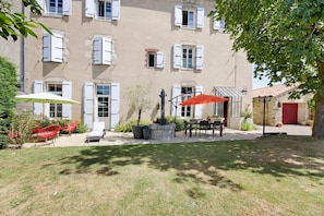 A building with multiple windows, a patio with outdoor furniture, and a tree providing shade.