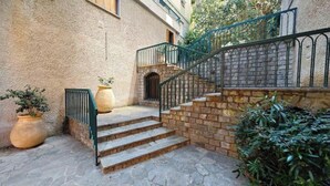Una escalera de piedra conduce a la entrada de un edificio, con barandilla de metal, plantas en macetas y un muro de ladrillo.