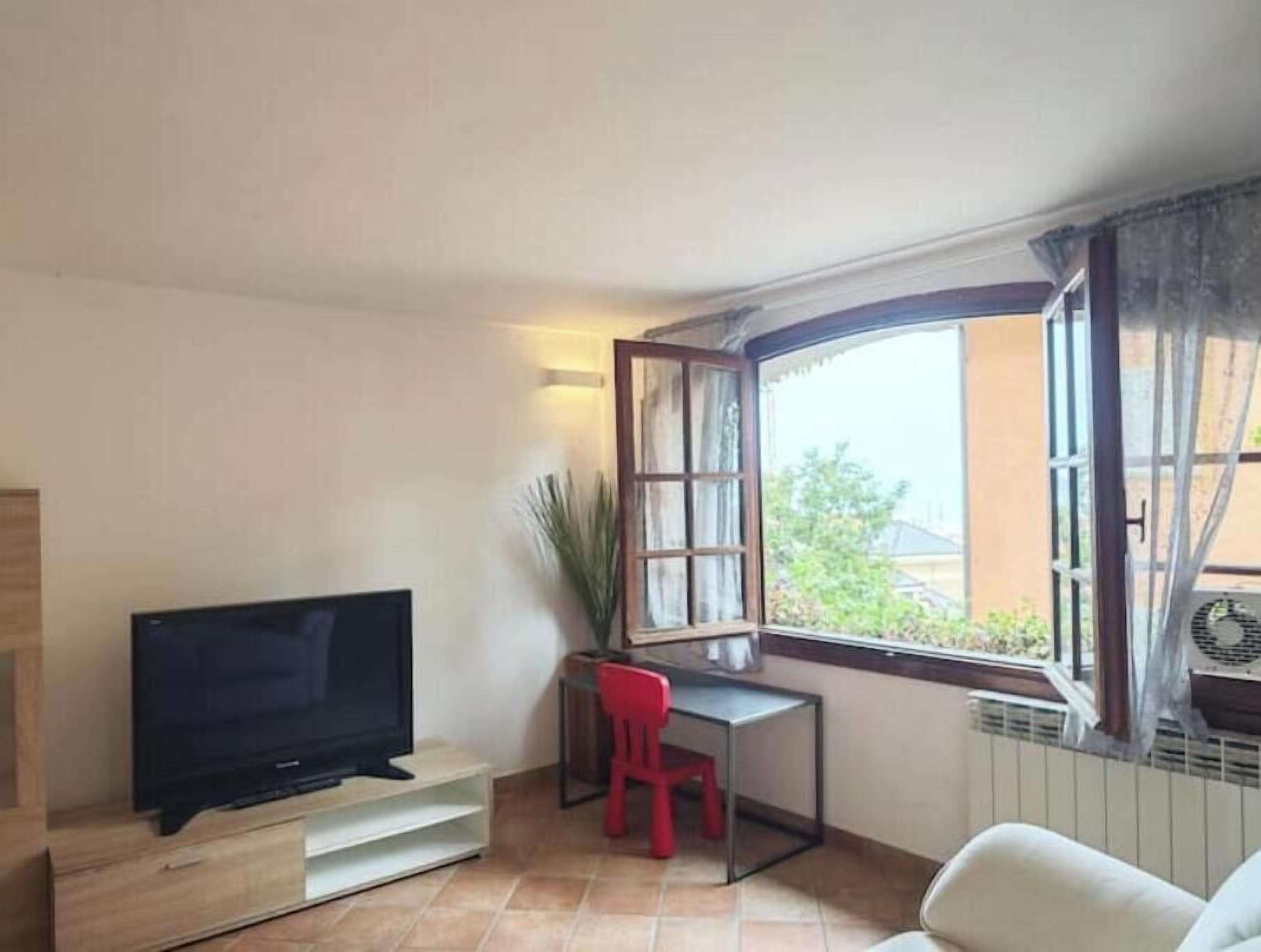 Una sala de estar con televisión, escritorio, una silla roja y una ventana con cortinas.