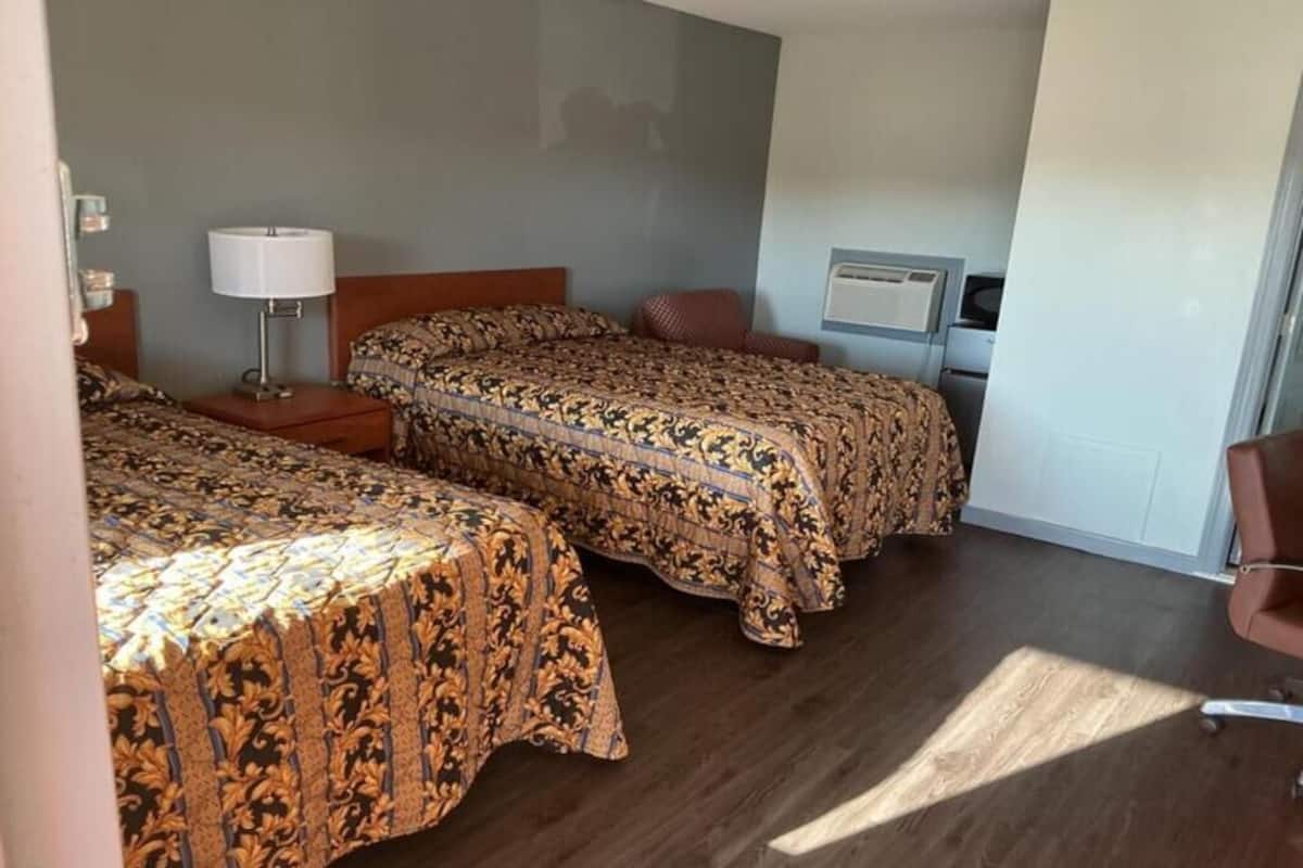 A hotel room with two beds, a nightstand with a lamp, a chair, and a wall-mounted air conditioner.