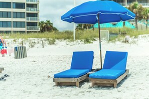 Zwei Holzliegen mit blauen Kissen unter einem blauen Sonnenschirm an einem Sandstrand.