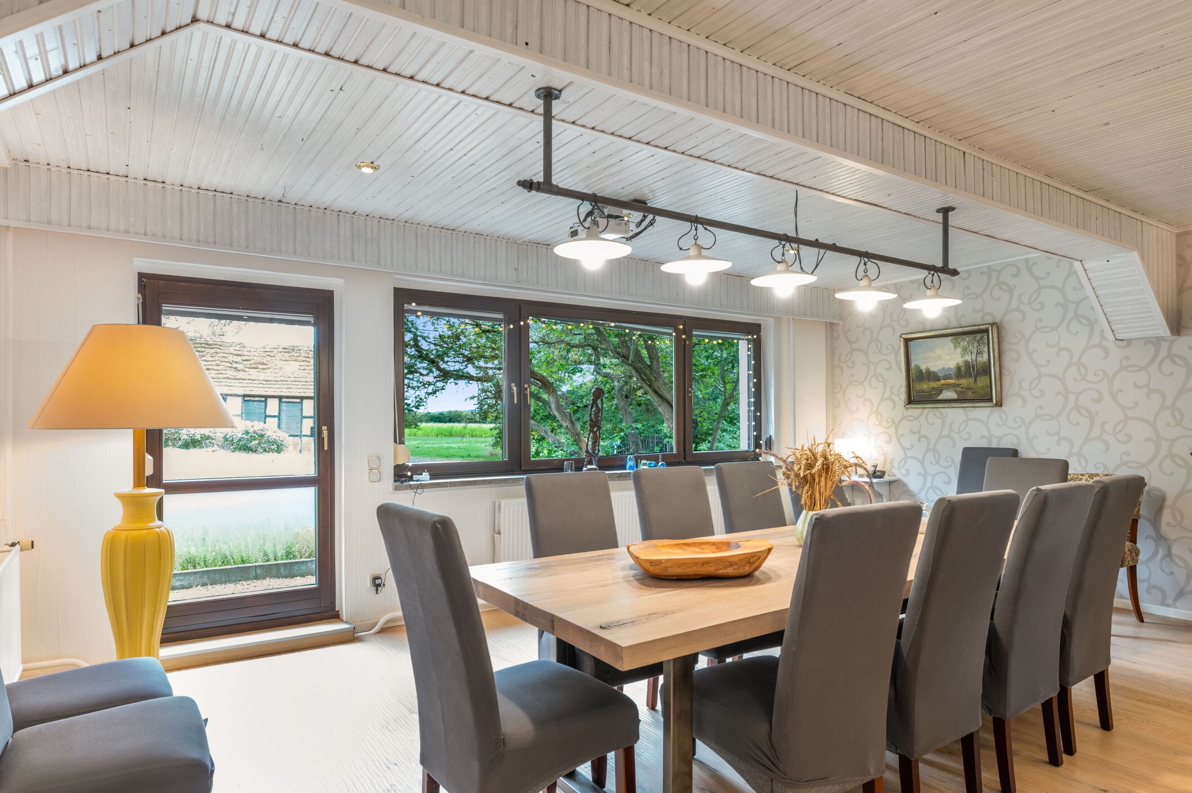 A dining area with a wooden table, grey chairs, a yellow lamp, and a view of greenery through large windows.