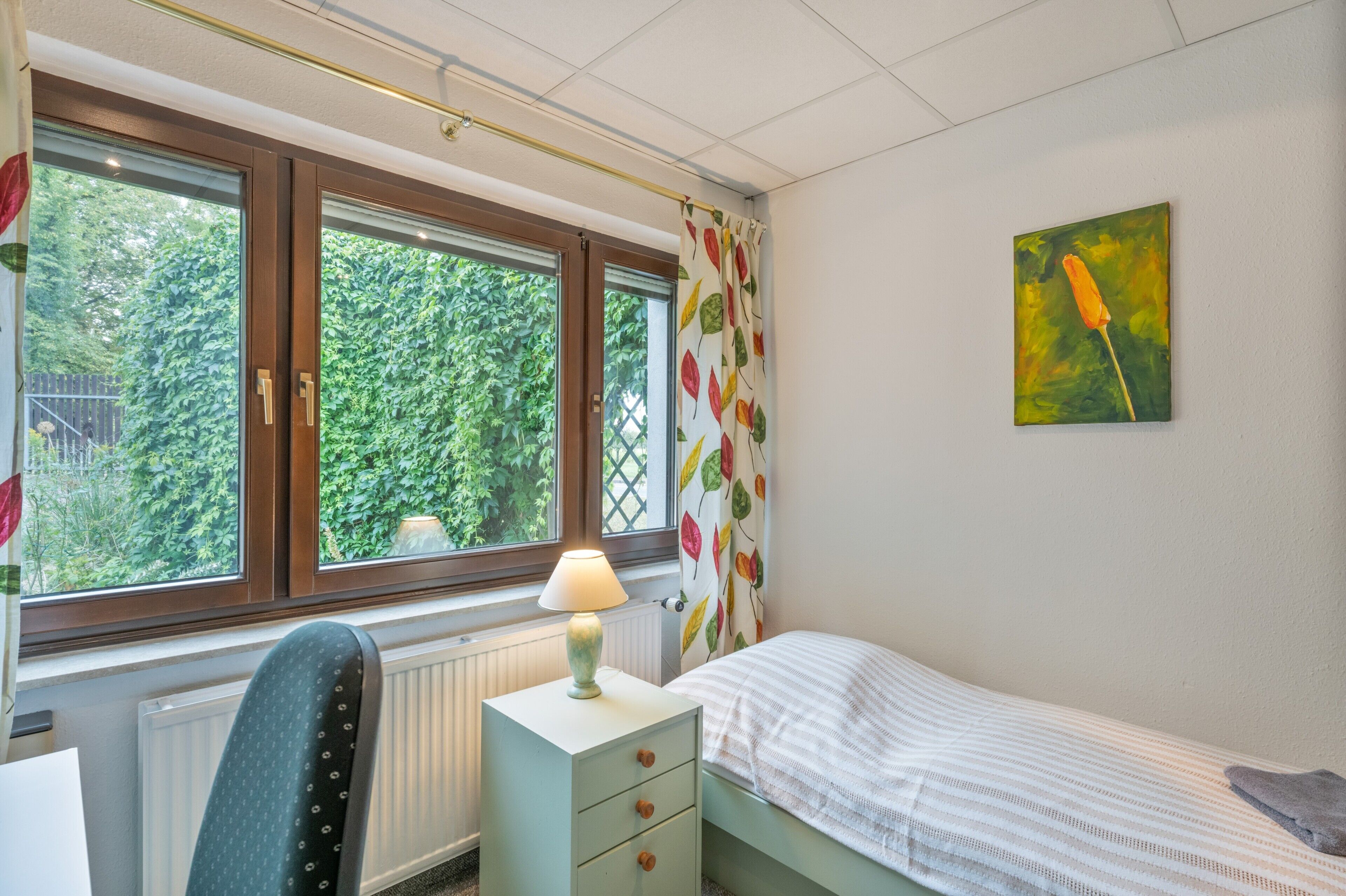 Ein Schlafzimmer mit Bett, Schreibtisch, Stuhl, Heizkörper und einem Fenster mit Vorhängen.