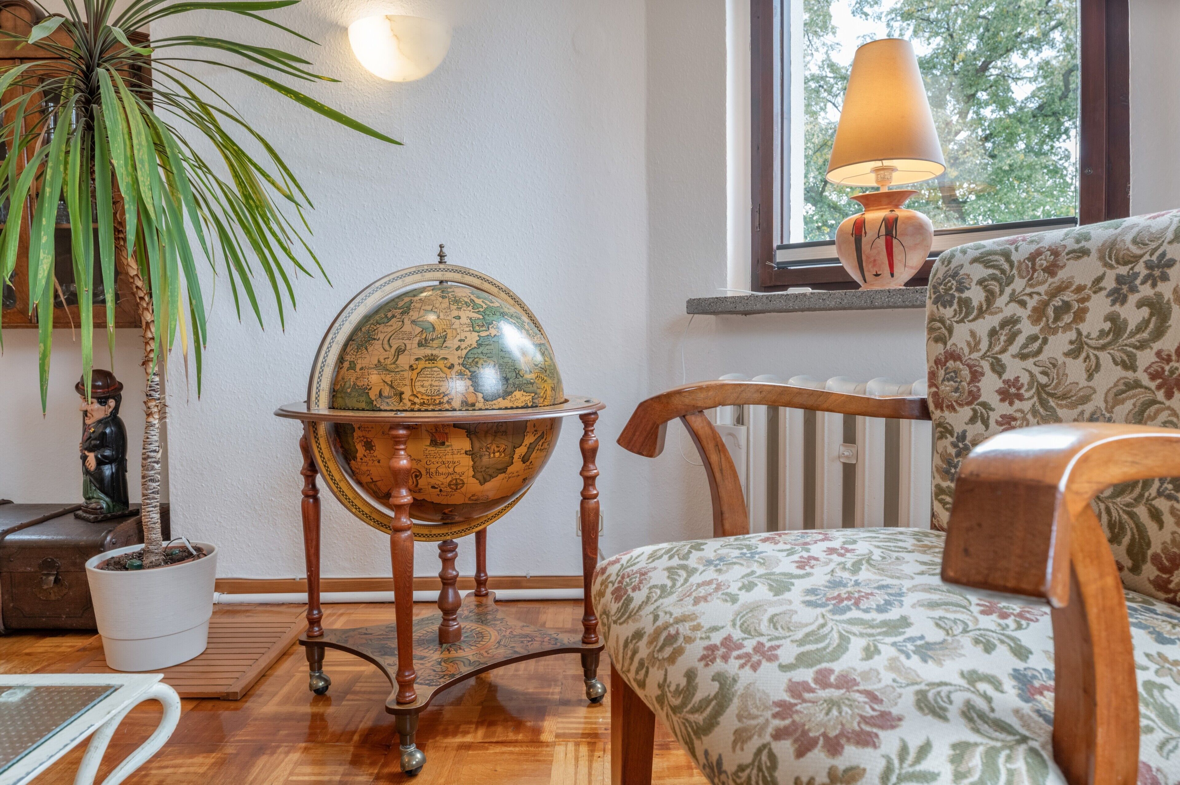 A room with a globe on a wooden stand, a potted plant, a wooden chair, and a lamp by a window.
