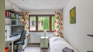 Un dormitorio con cama, escritorio con computadora, silla, estantería y ventana con cortinas.