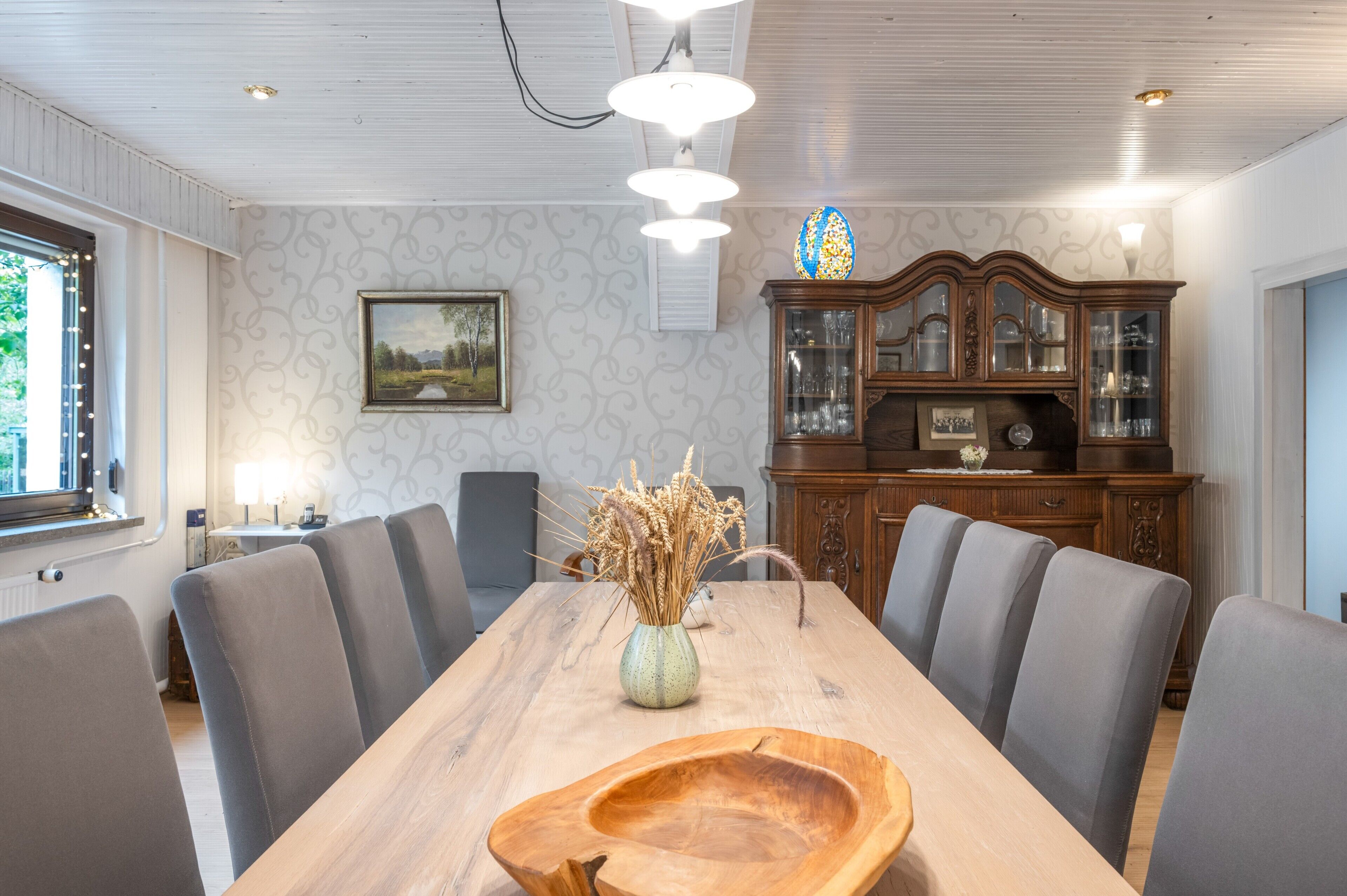 Ein Essbereich mit einem langen Holztisch, grau gepolsterten Stühlen, einer Vase mit Trockenpflanzen und einer Holztruhe mit Glastüren.